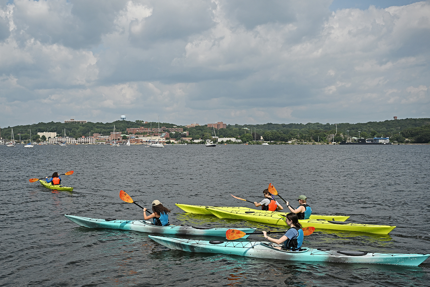 Students in kayaks paddle off under a cloudy sky.