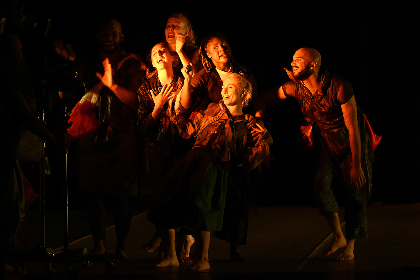 Modern dancers perform in a tight group under a harsh yellow light coming from the side of the stage.